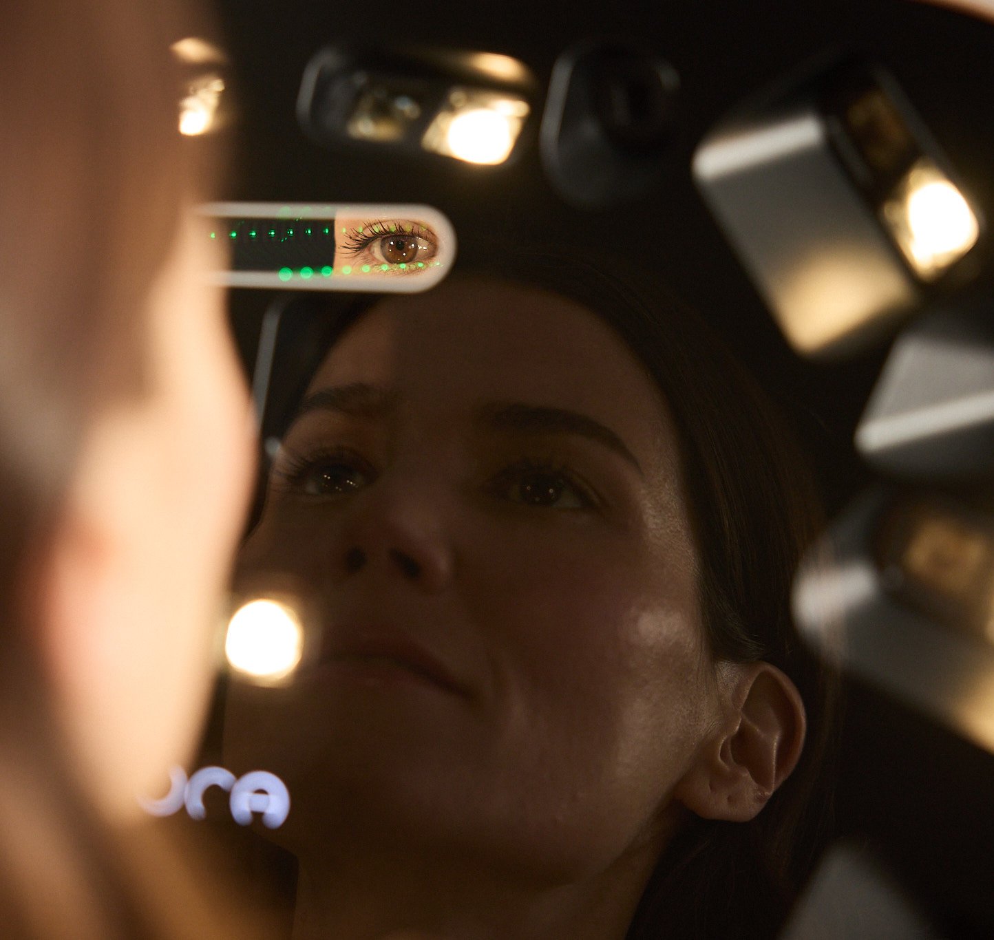 Close-up of a woman's eye reflected in a small mirror-like instrument with green LED indicators and warm lighting in the background.